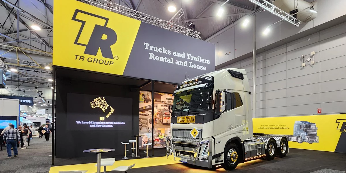 TR Group exhibition booth at 2023 Brisbane Truck Show, featuring a large display with the TR Group logo, slogan 'Trucks and Trailers Rental and Lease,' and a white road train truck on display. The booth includes a seating area and digital screens showcasing branch locations across Australia and New Zealand.